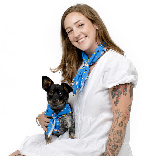 A woman with tattoos wearing a white dress and a blue patterned scarf, holding a small black and gray dog that also wears a matching blue patterned scarf, against a solid white background.