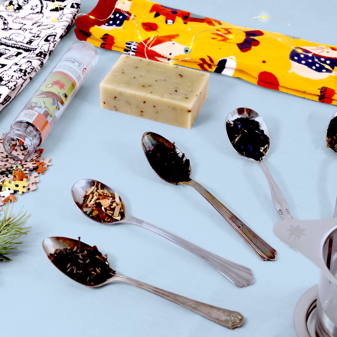 Product flatlay on a light blue background featuring different tea samples displayed on silver spoons, a bar of soap with visible herbs or tea leaves, decorative socks in black/white and yellow patterns, a small bottle of puzzle pieces, and pine sprigs decorated with tiny lights.