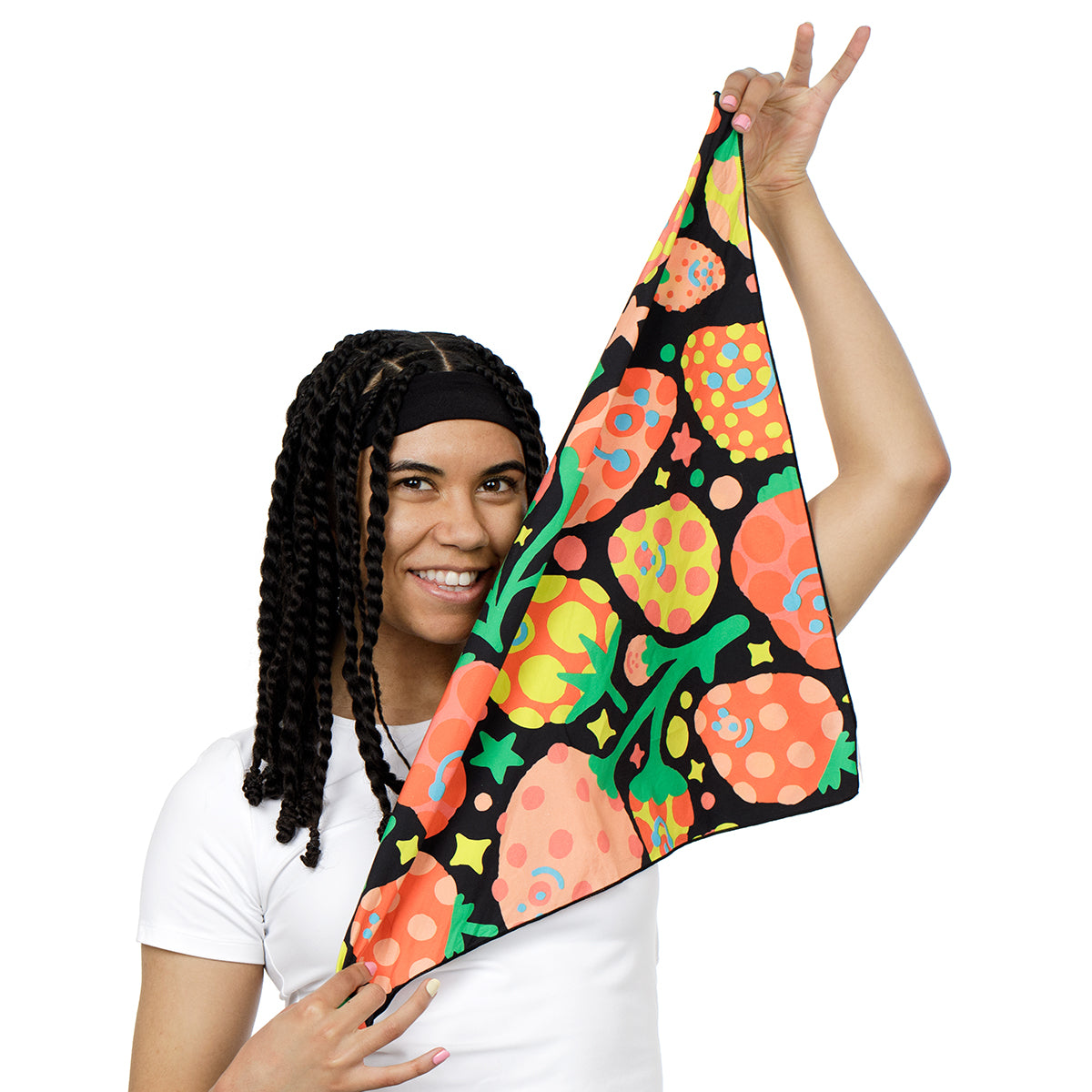 A woman with long black braids wearing a white t-shirt holds up a black bandana featuring a vibrant, colorful pattern of orange, yellow, and green tropical fruits on a solid white background.
