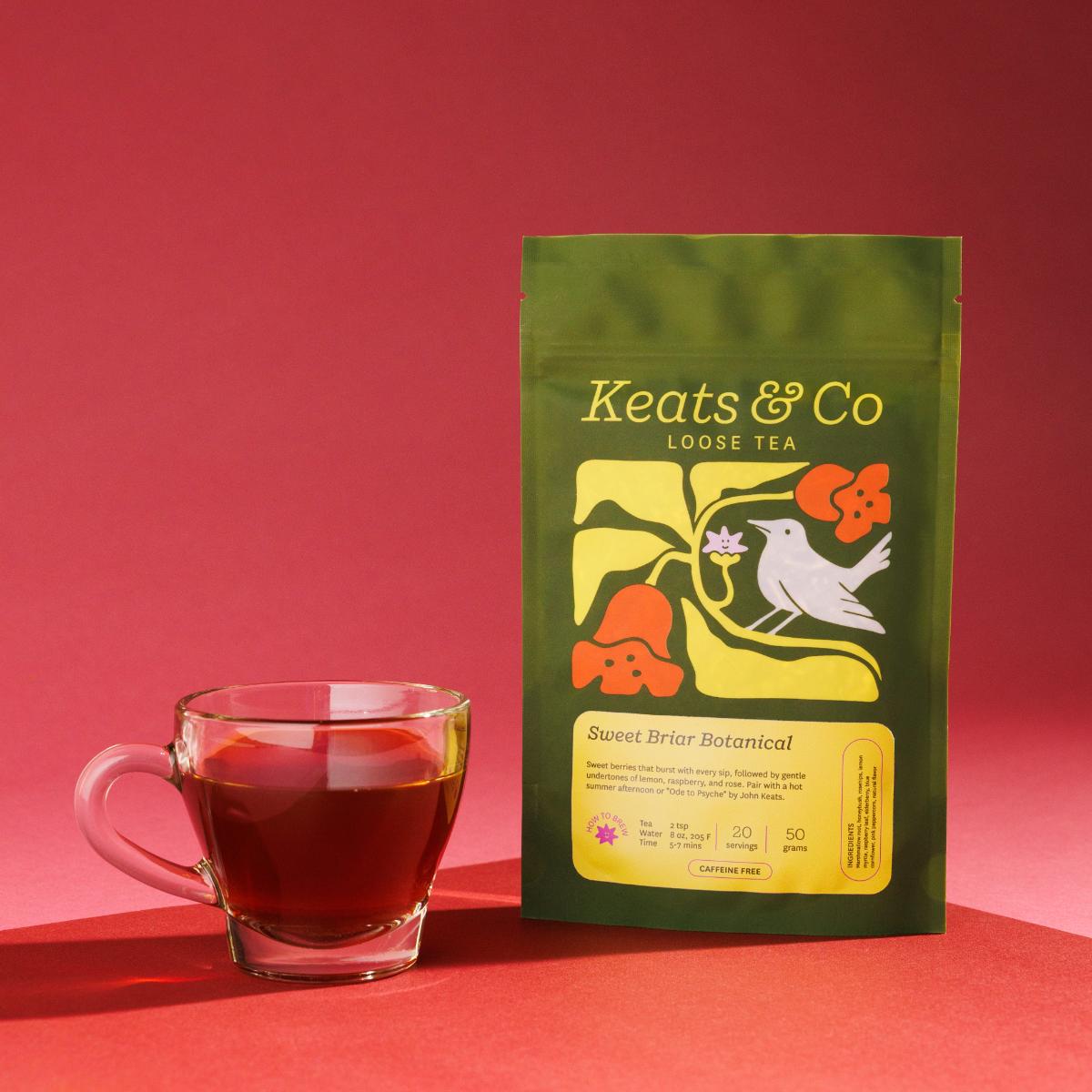 Green pouch of Keats & Co Loose Tea Sweet Briar Botanical against a pink background, featuring illustrated artwork of a white bird and orange flowers, displayed next to a clear glass mug filled with red-ish brewed tea on a pink surface.