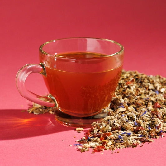 Glass cup of orange-ish Sweet Briar Botanical tea positioned next to a pile of loose herbals on a pink backround. A Good Store product.