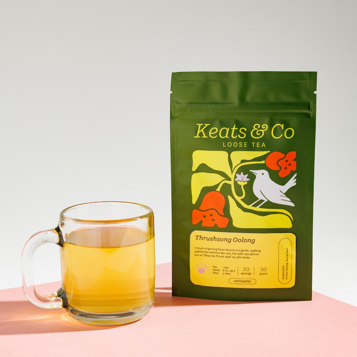Green pouch of Keats & Co Loose Tea Thursong Oolong against a white background, featuring illustrated artwork of a white bird and orange flowers, displayed next to a clear glass mug filled with yellowish brewed tea on a pink surface.