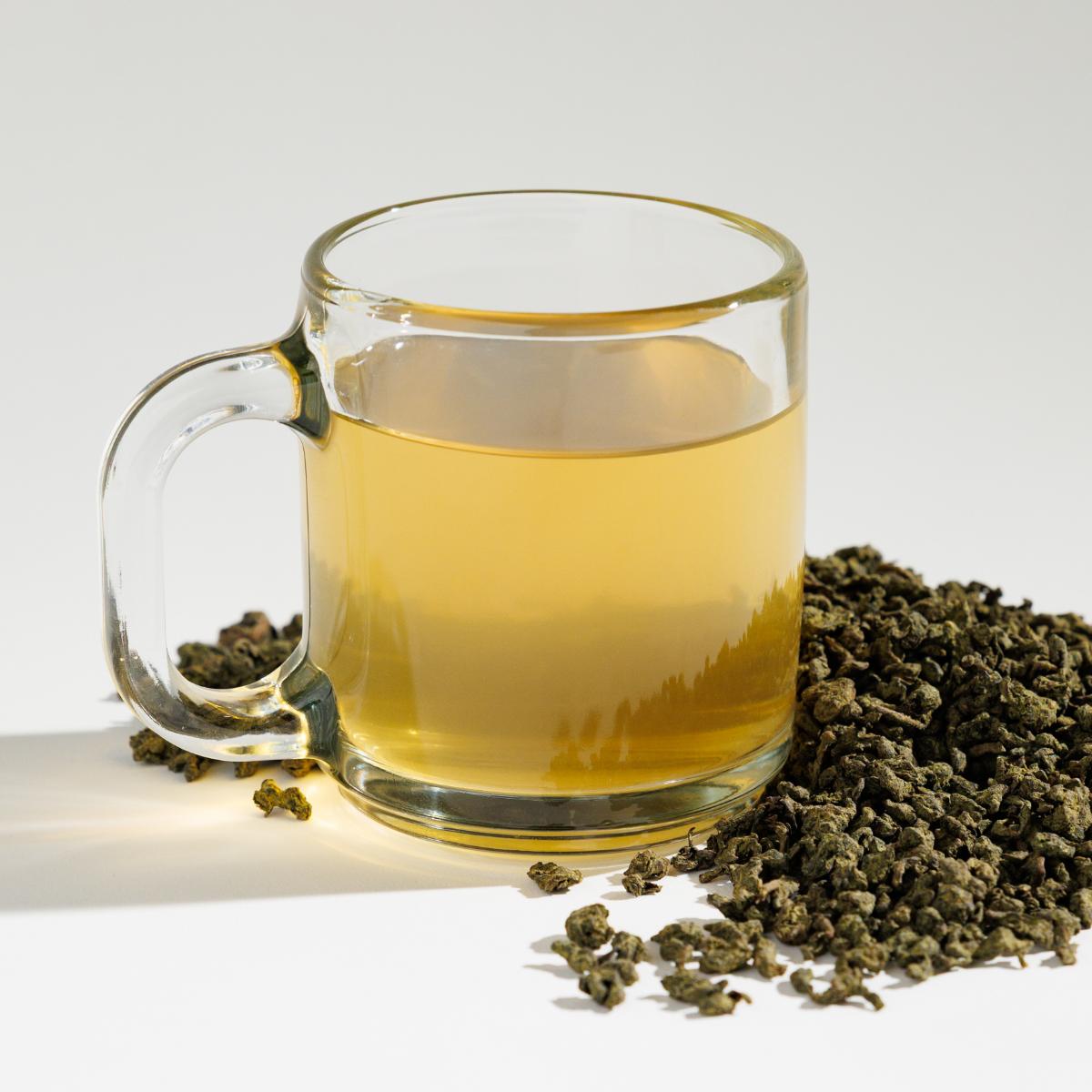 Glass cup of yellowish Thrushsong Oolong tea positioned next to a pile of loose leaf tea blend containing rolled Oonlong tea leaves on a reflective white surface against a soft white background. A Good Store product.