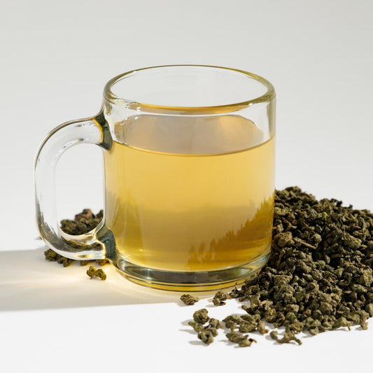 Glass cup of yellowish Thrushsong Oolong tea positioned next to a pile of loose leaf tea blend containing rolled Oonlong tea leaves on a reflective white surface against a soft white background. A Good Store product.