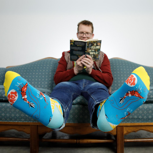 Photo of Hank Green wearing Awesome Socks sitting on a couch reading a book. A Good Store product.