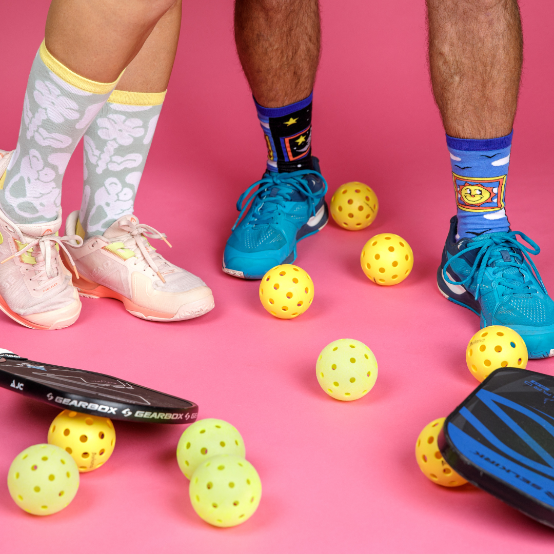 Two pairs of legs wearing Awesome Sock Club crew socks with different patterns. The people are wearing tennis shoes and are surrounded by pickleball equipment. A Good Store product. 