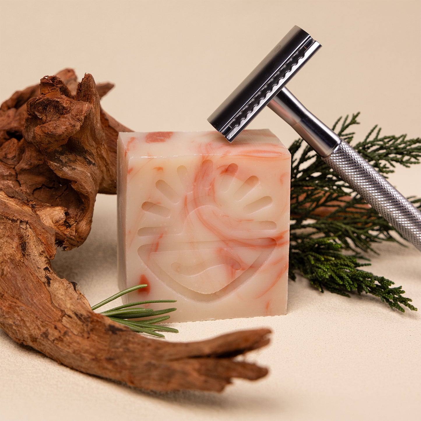 A marbled cream and pink shaving soap bar with a carved sun face design, styled with a silver safety razor, pine branches, and driftwood on a beige background.