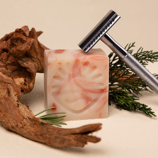 A marbled cream and pink shaving soap bar with a carved sun face design, styled with a silver safety razor, pine branches, and driftwood on a beige background.