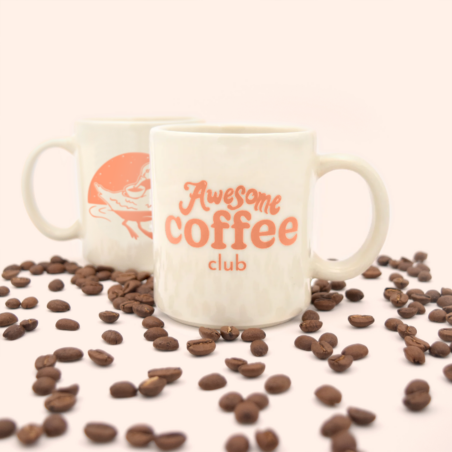 White cafe mug with the Awesome Coffee Club logo in orange written on the front. Behind the mug is a second mug with the outline of a duck drinking out of a cup, showing the back of the mug. Coffee beans surround both mugs. A Good Store subscription product. 