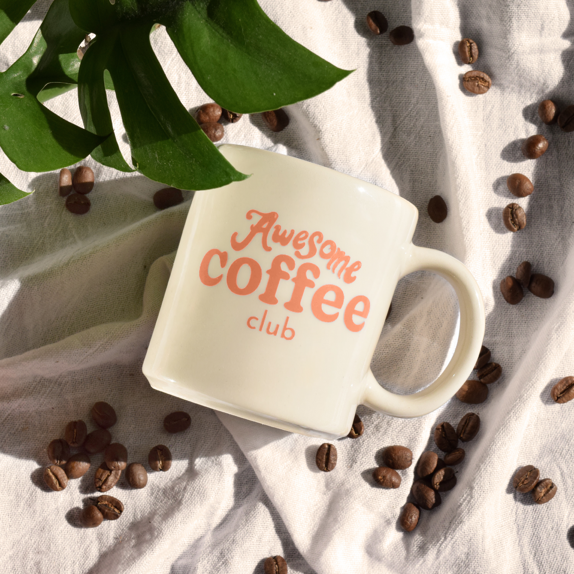 White cafe mug with "Awesome Coffee Club" written in orange. The mug is laid on a sheet with a plant in the corner and coffee beans surrounding it.