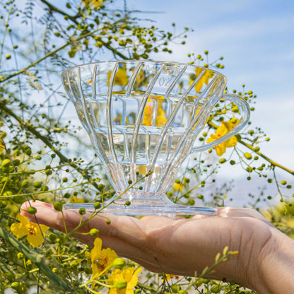 Clear, plastic coffee pour over being held in a hand with yellow flowers and sky in the background. Available through the Awesome Coffee Club. A Good Store subscription product.