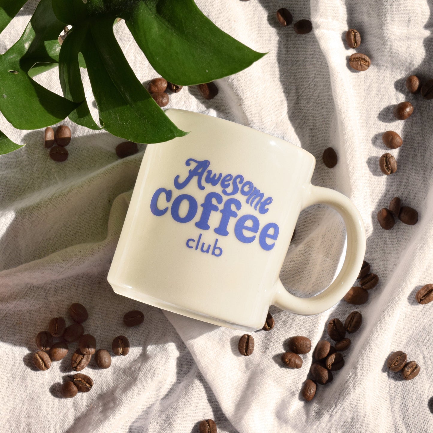 White cafe mug with "Awesome Coffee Club" written in purple. The mug is laid on a sheet with a plant in the corner and coffee beans surrounding it.